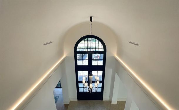 Looking down from a high ceiling hallway toward a grand arched black entry door with a chandelier